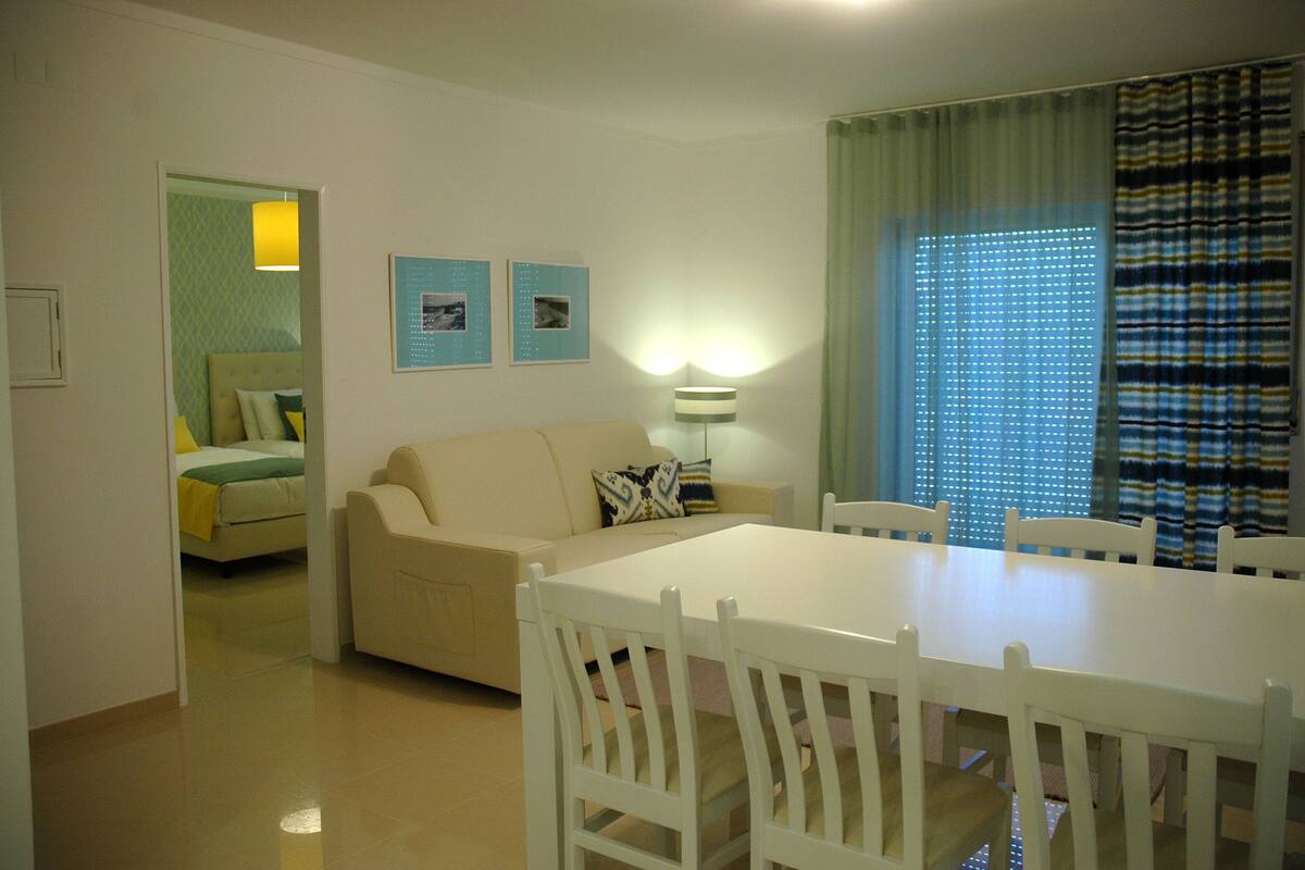 Una sala de estar con un sofá beige, una mesa de comedor con sillas blancas y una cama al fondo.