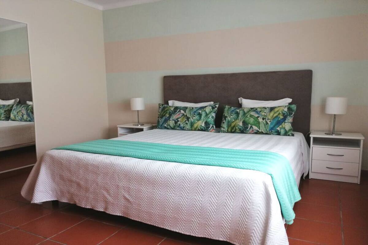 Una cama bien hecha con colcha blanca y verde, dos mesitas de noche con lámparas y un espejo en la pared.