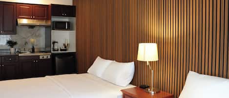 A hotel room with two beds, a wooden panel wall, a kitchenette, and a nightstand with a lamp.