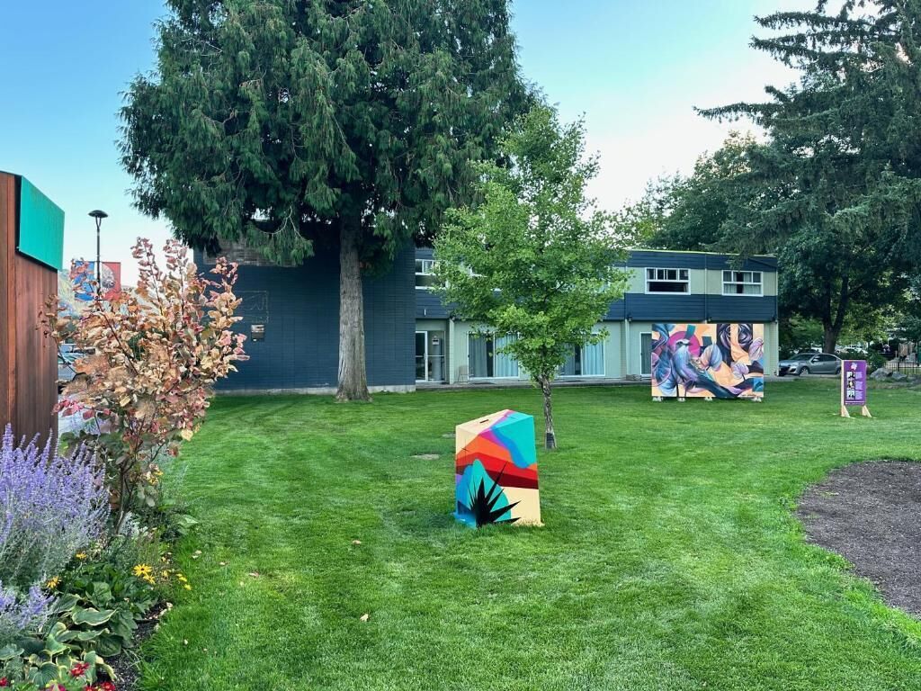 Un espacio con césped, una escultura colorida, un árbol y un edificio con un mural al fondo.