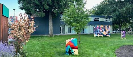 Un espacio con césped, una escultura colorida, un árbol y un edificio con un mural al fondo.