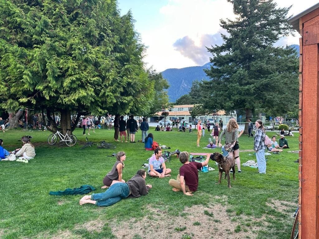 An outdoor gathering with people sitting on grass, trees, and a building in the background.
