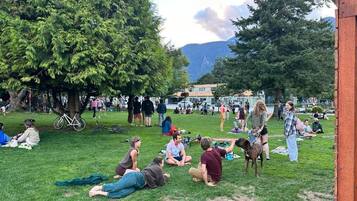 An outdoor gathering with people sitting on grass, trees, and a building in the background.