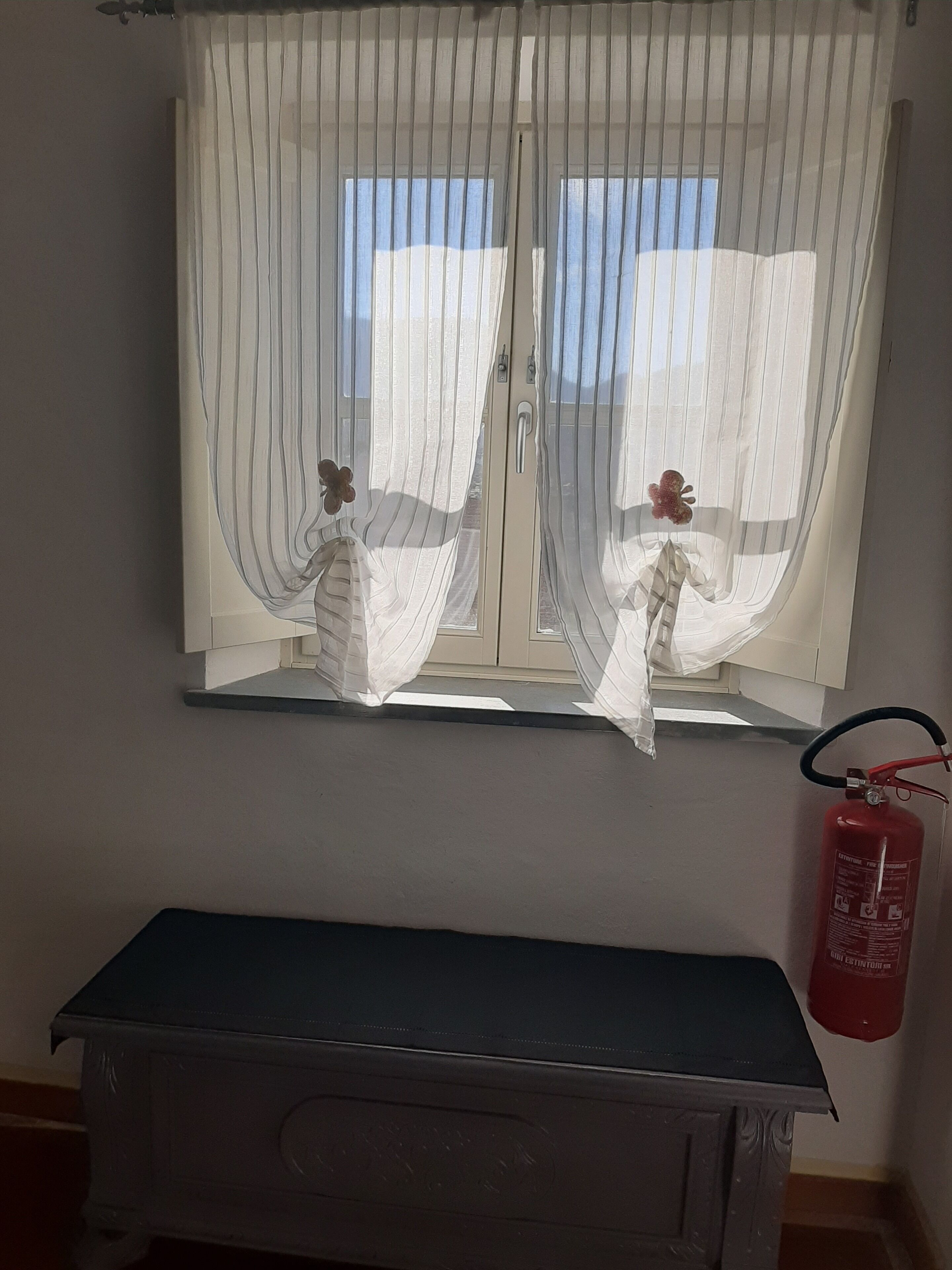 A room with a window covered by sheer curtains, a fire extinguisher mounted on the wall, and a bench in the foreground.