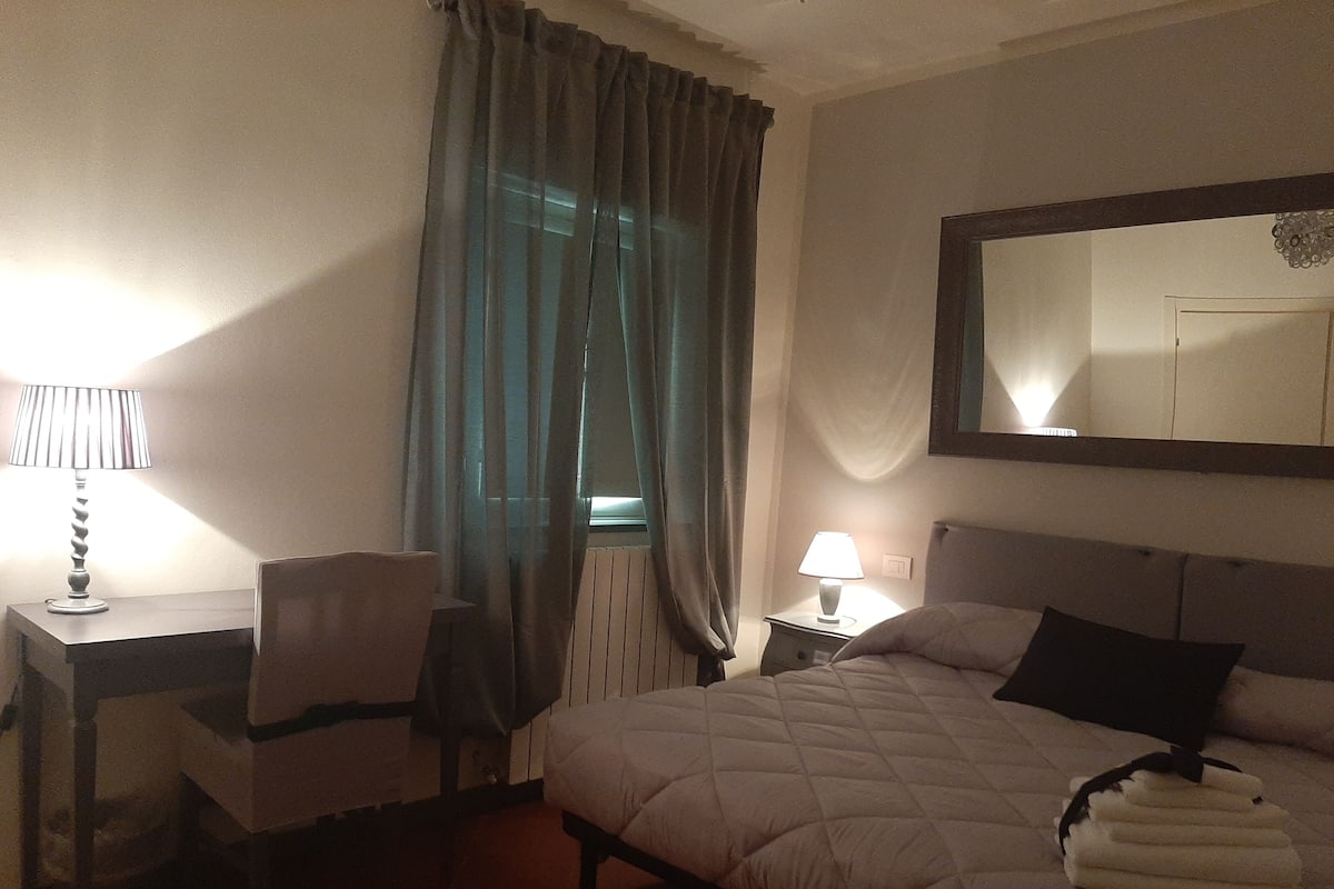 Quarto de hotel com cama, escrivaninha, cadeira, abajur e janela com cortinas.