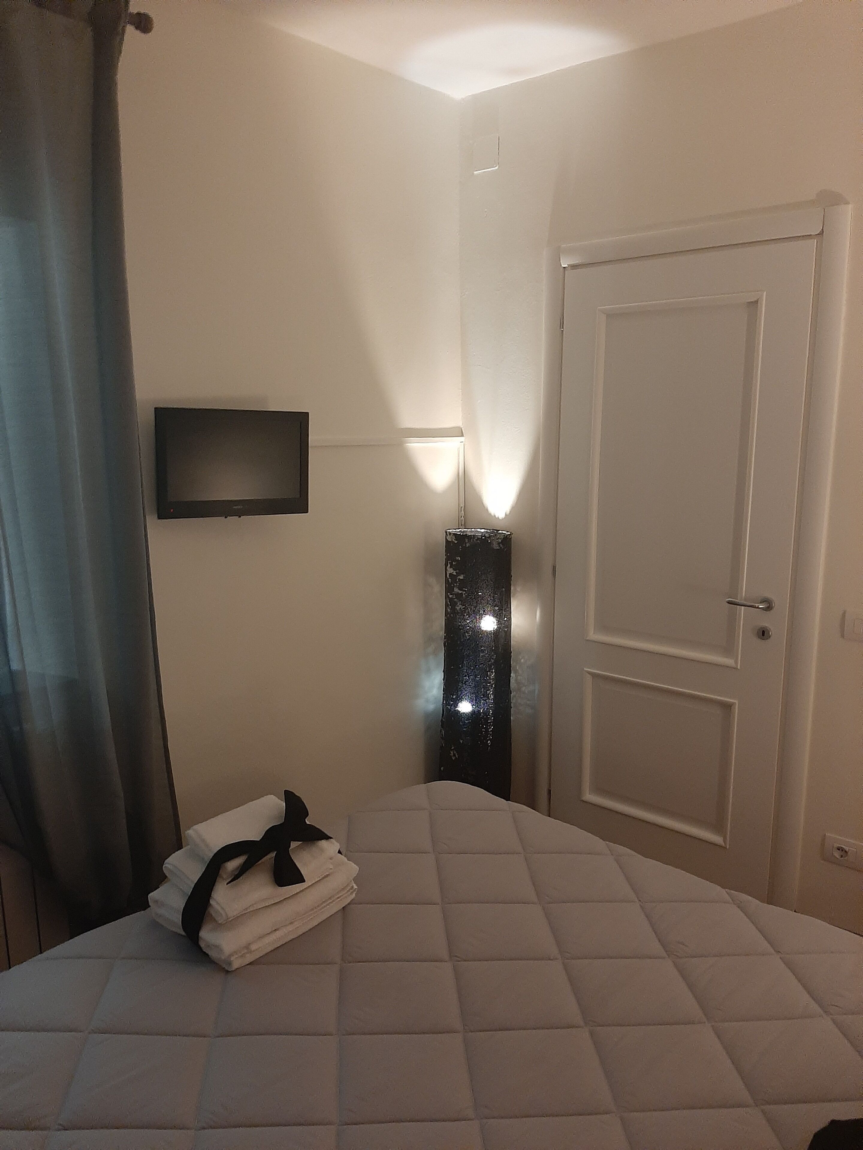A hotel room with a bed, a TV mounted on the wall, a door, and a wall-mounted light fixture.