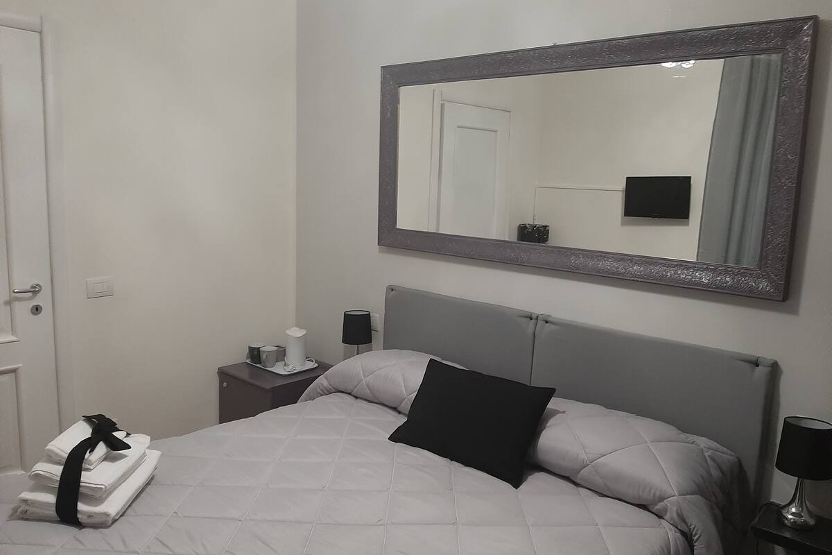 Quarto de hotel com cama, mesa de cabeceira, abajur, espelho e porta.