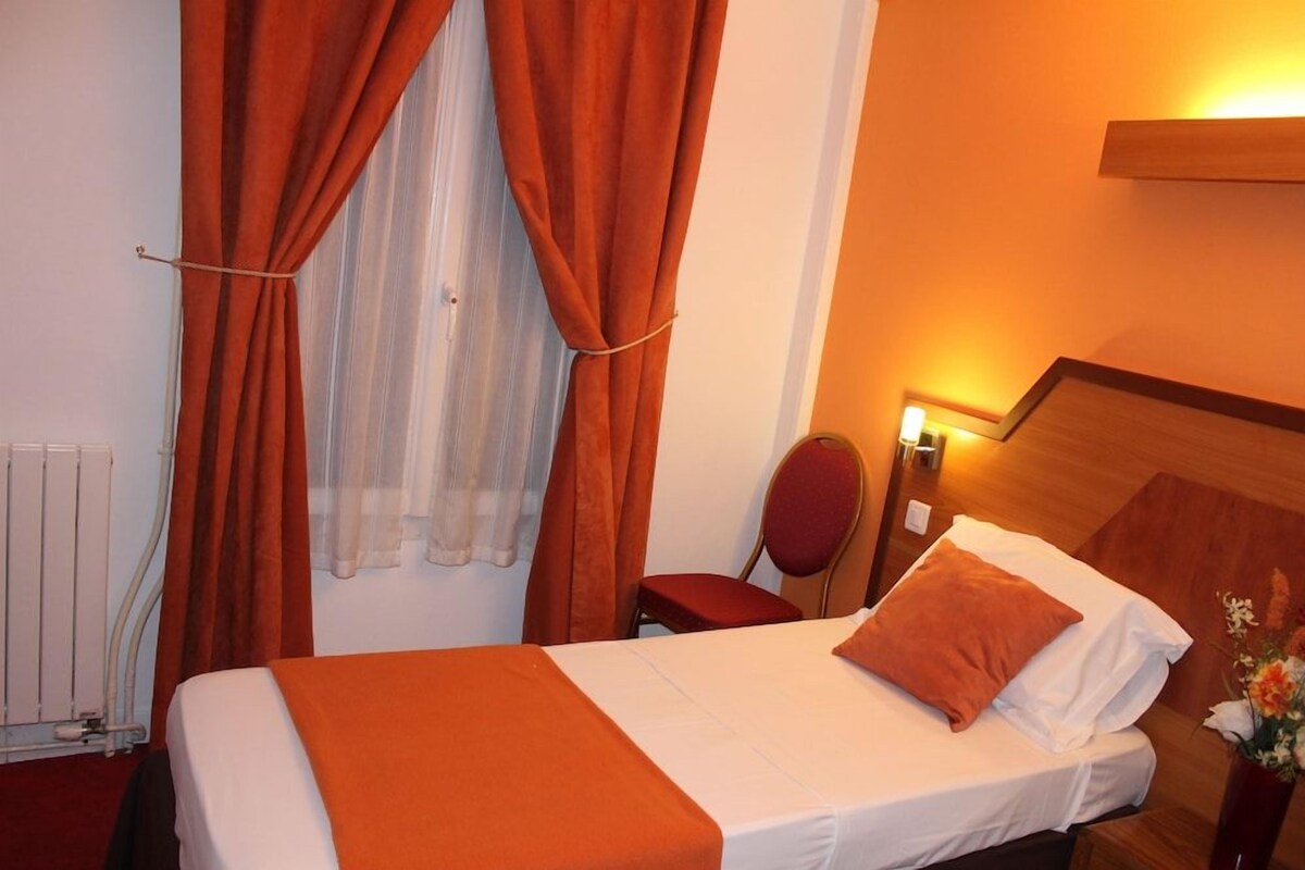 Une chambre d’hôtel avec un lit, des rideaux orange, une chaise, une table de chevet avec une lampe et un radiateur.