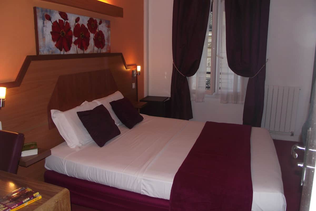 Een hotelkamer met een houten hoofdeinde, een groot bed met wit en bordeauxrood beddengoed, een schilderij van rode bloemen aan de muur en een raam met donkere gordijnen.