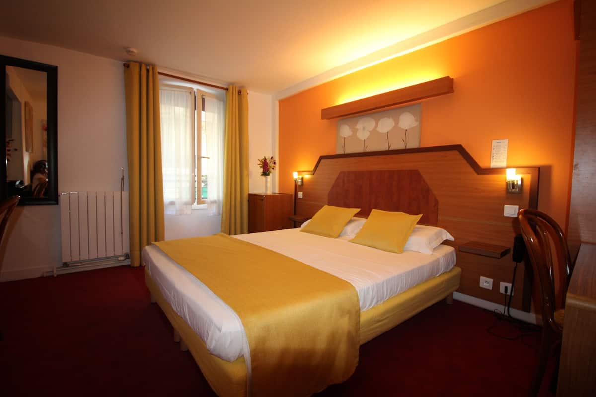 Een hotelkamer met een groot bed, een houten hoofdbord, een spiegel, een radiator en een raam met gordijnen.