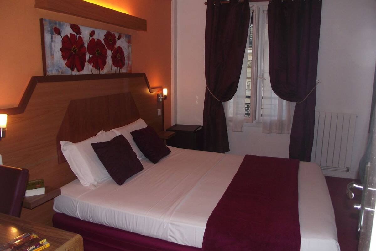 Een hotelkamer met een bed, nachtkastje, schilderij, raam met gordijnen en een radiator.