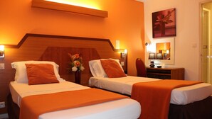 Een hotelkamer met twee eenpersoonsbedden, elk met een oranje sprei en kussens. Er is een houten hoofdbord, een klein tafeltje met een bloemenvaas, en een spiegel aan de muur.