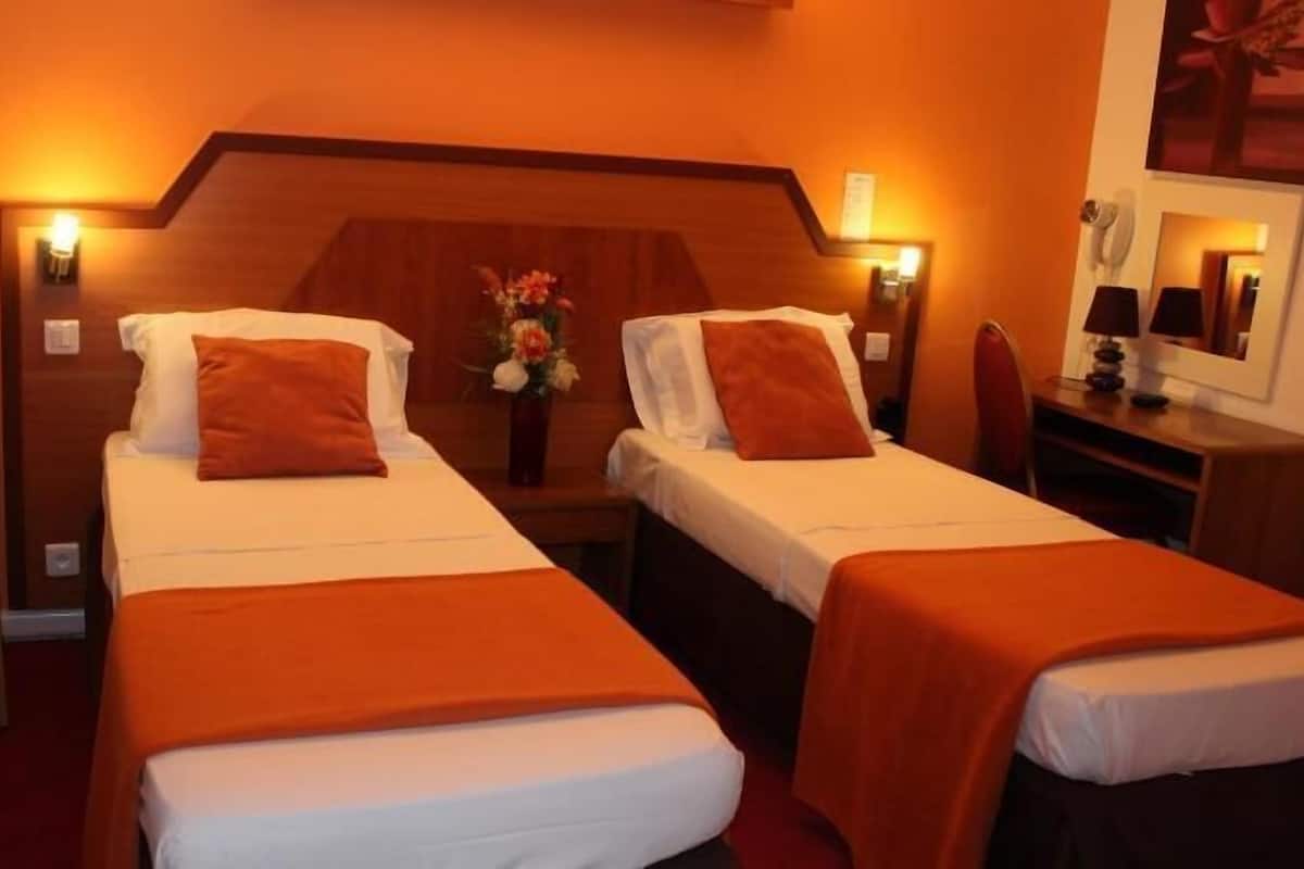 Een hotelkamer met twee éénpersoonsbedden, een houten hoofdbord, een tafeltje met een bloemenvaas, een stoel en een bureau met een lamp.