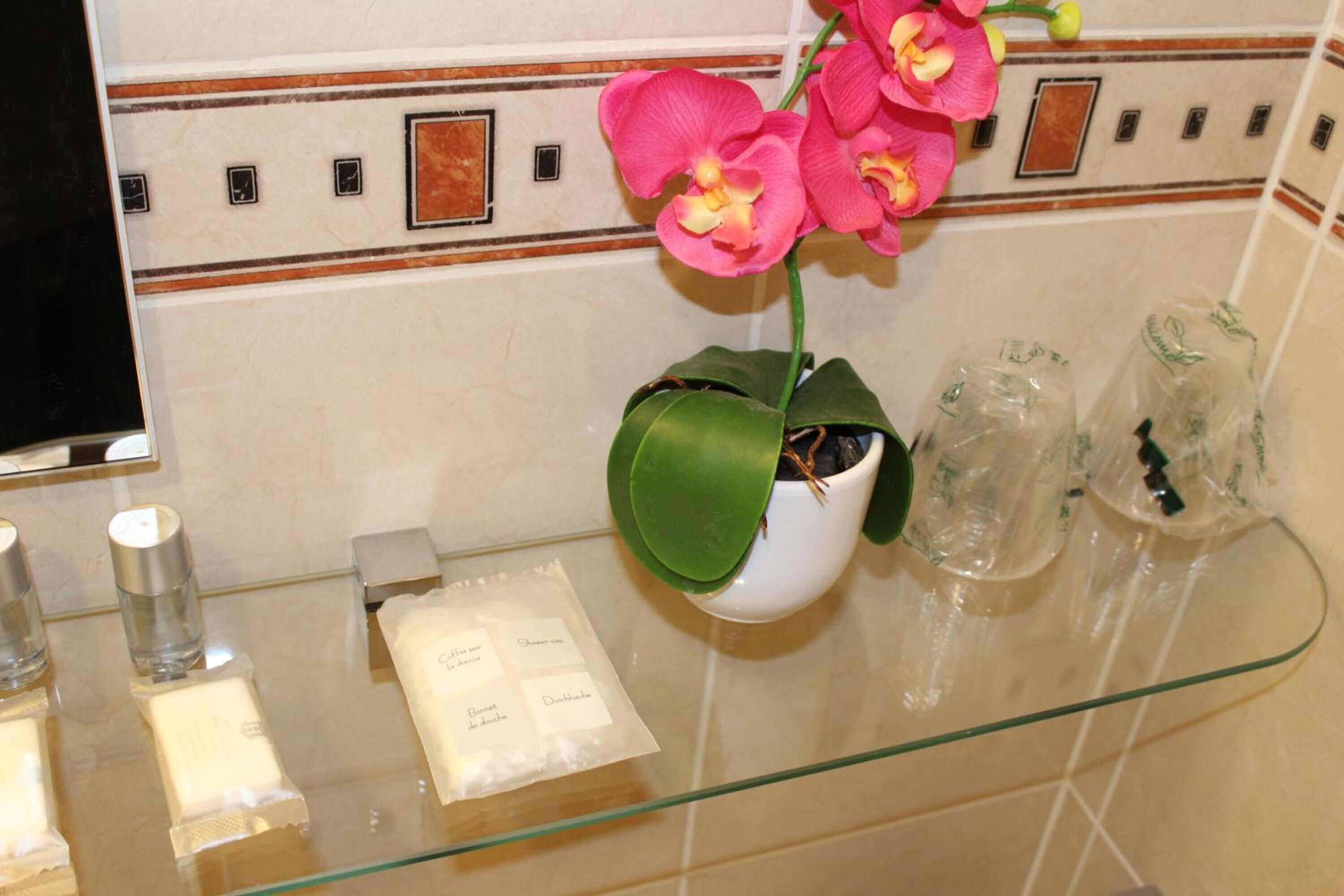 A bathroom with a glass shelf, a white vase with pink orchids, and a mirror.