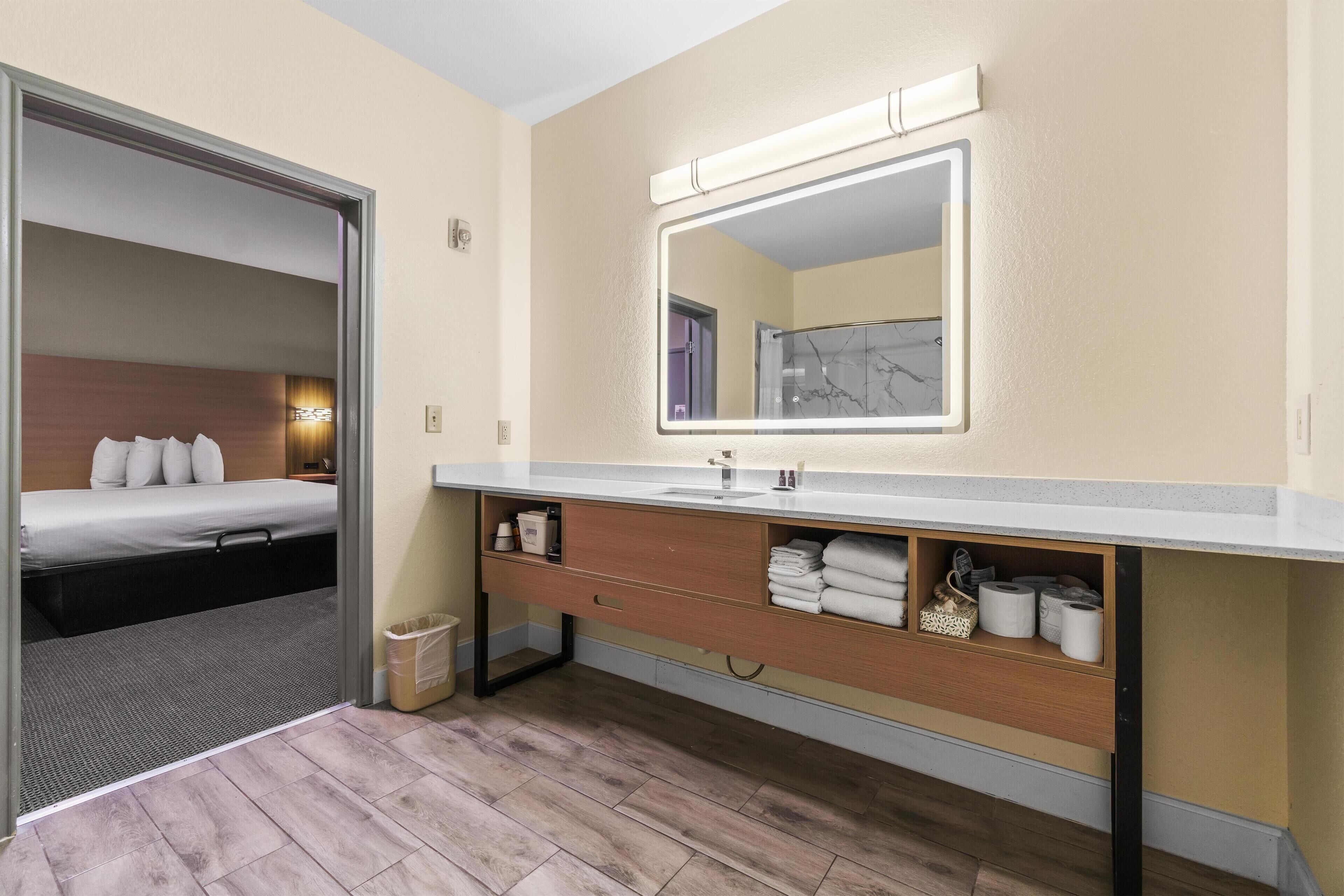 Hotellihuone, jossa on puulattia, valkoisilla vuodevaatteilla varustettu sänky, suuri peili kylpyhuoneessa sekä pesuallas, jossa on pyyhkeitä ja hygieniatuotteita.