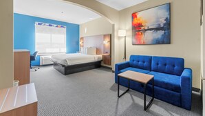 A hotel room with a bed, a blue sofa, a desk, and a painting on the wall.