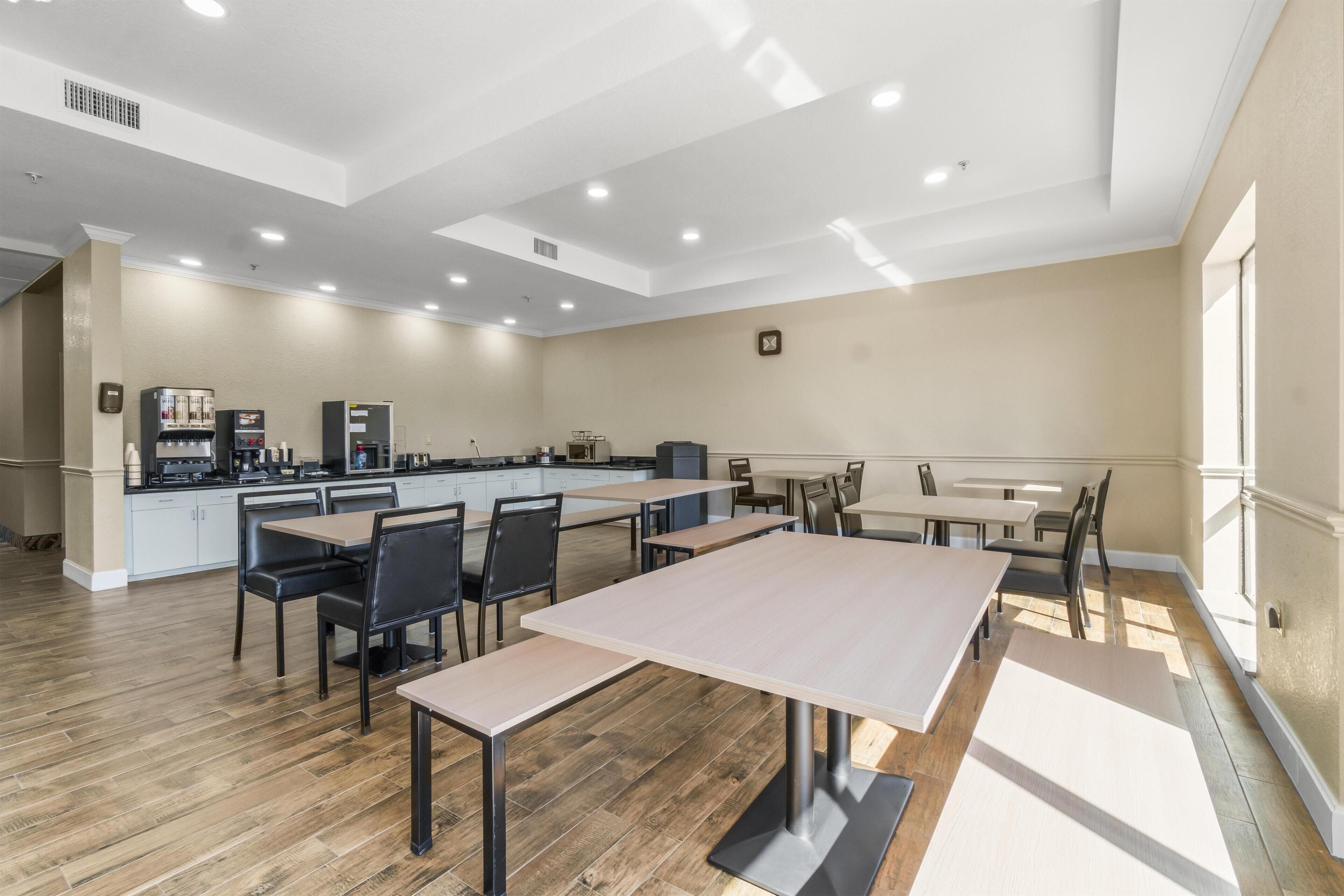 Un comedor amplio con varias mesas y sillas, una kitchenette al fondo y un ventanal que proporciona luz natural.