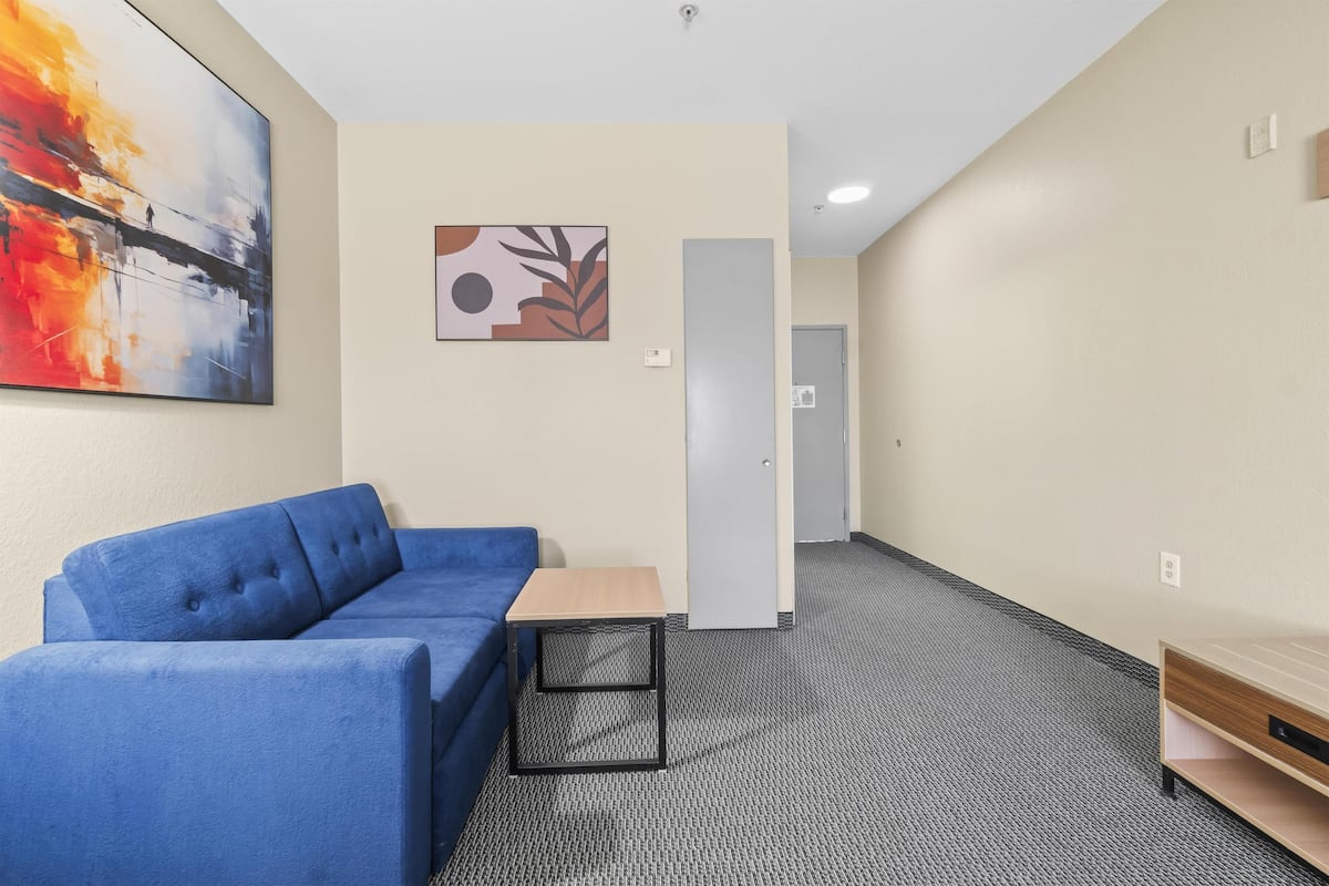 A modern hotel room with a blue sofa, a small wooden table, and a grey carpet.