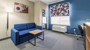 A hotel room with a blue sofa, a small table, a lamp, and abstract wall art.
