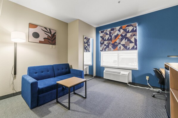 A hotel room with a blue sofa, a small table, a lamp, and abstract wall art.