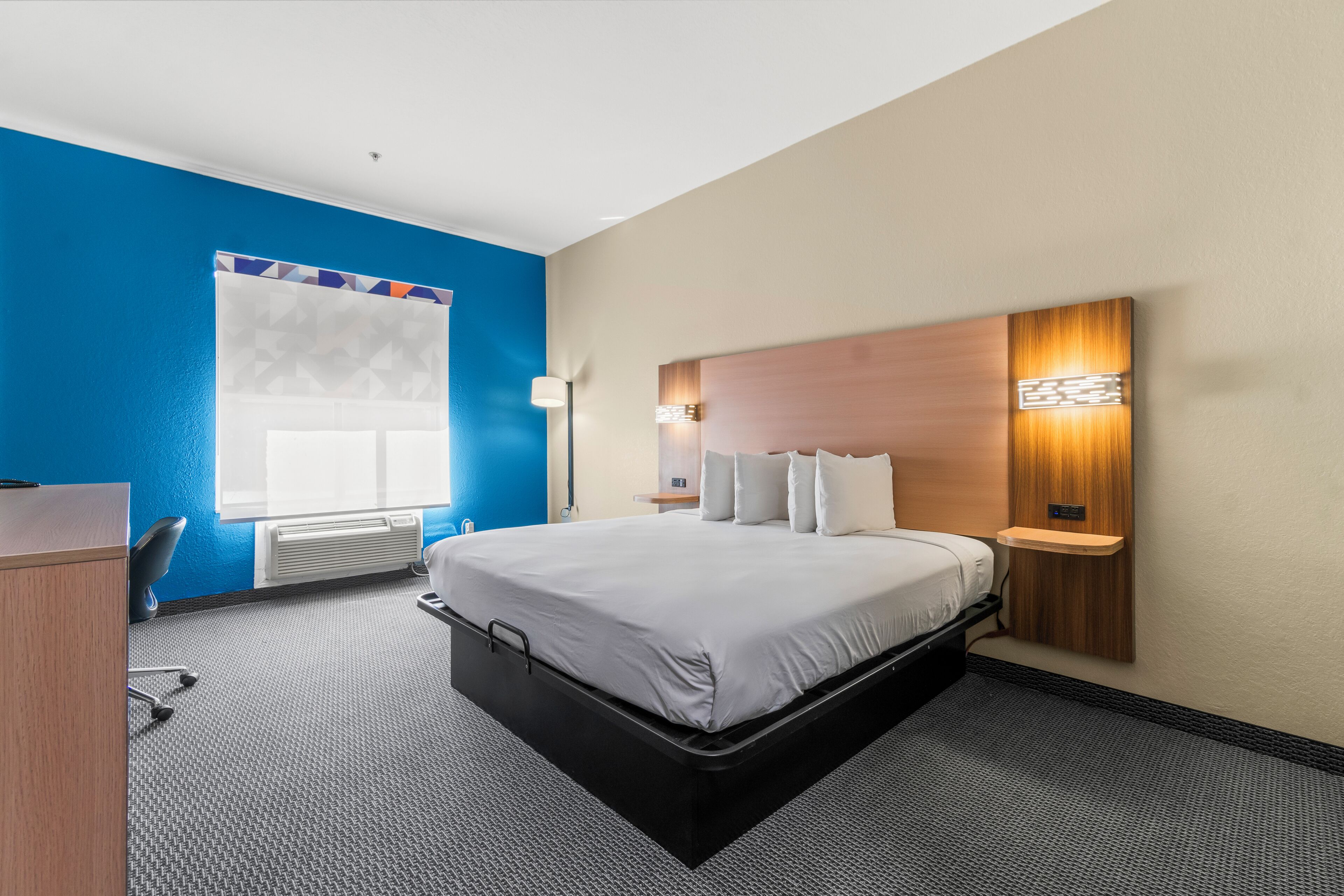 A hotel room with a bed, a desk, a chair, a wall-mounted shelf, and a window with blinds.