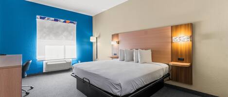 A hotel room with a bed, a desk, a chair, a wall-mounted shelf, and a window with blinds.