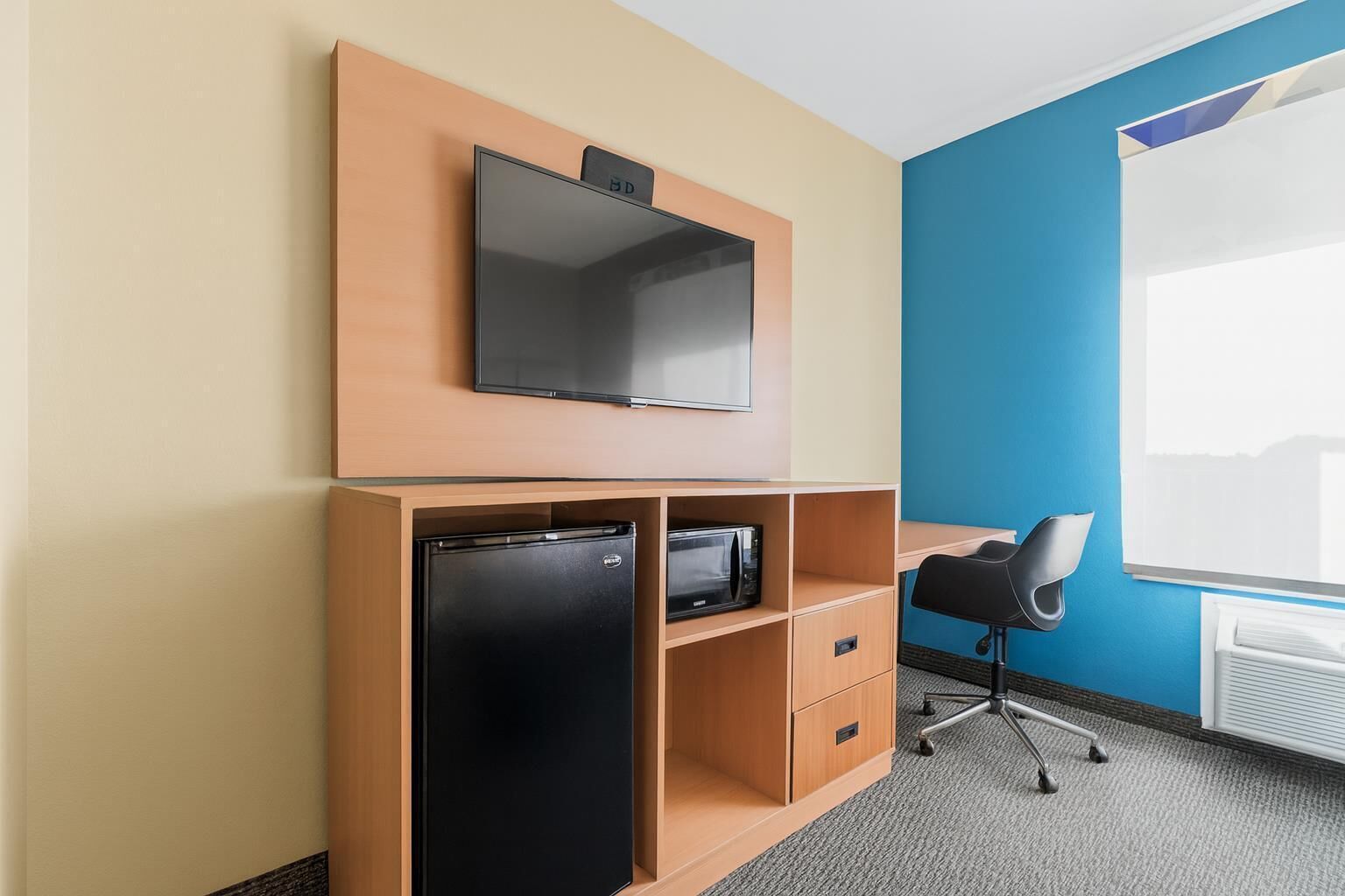 Habitación de hotel con televisión de pantalla plana montada en un panel de madera, refrigerador negro, microondas, escritorio con silla y ventana con vista.
