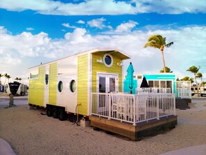 A small, yellow and white building with a balcony and a small porch, surrounded by palm trees and other structures.