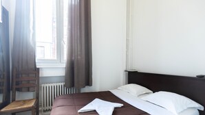 Ein ordentlich bezogenes Bett mit brauner Tagesdecke und weißen Kissen, ein Holzstuhl und ein Fenster mit Vorhängen.