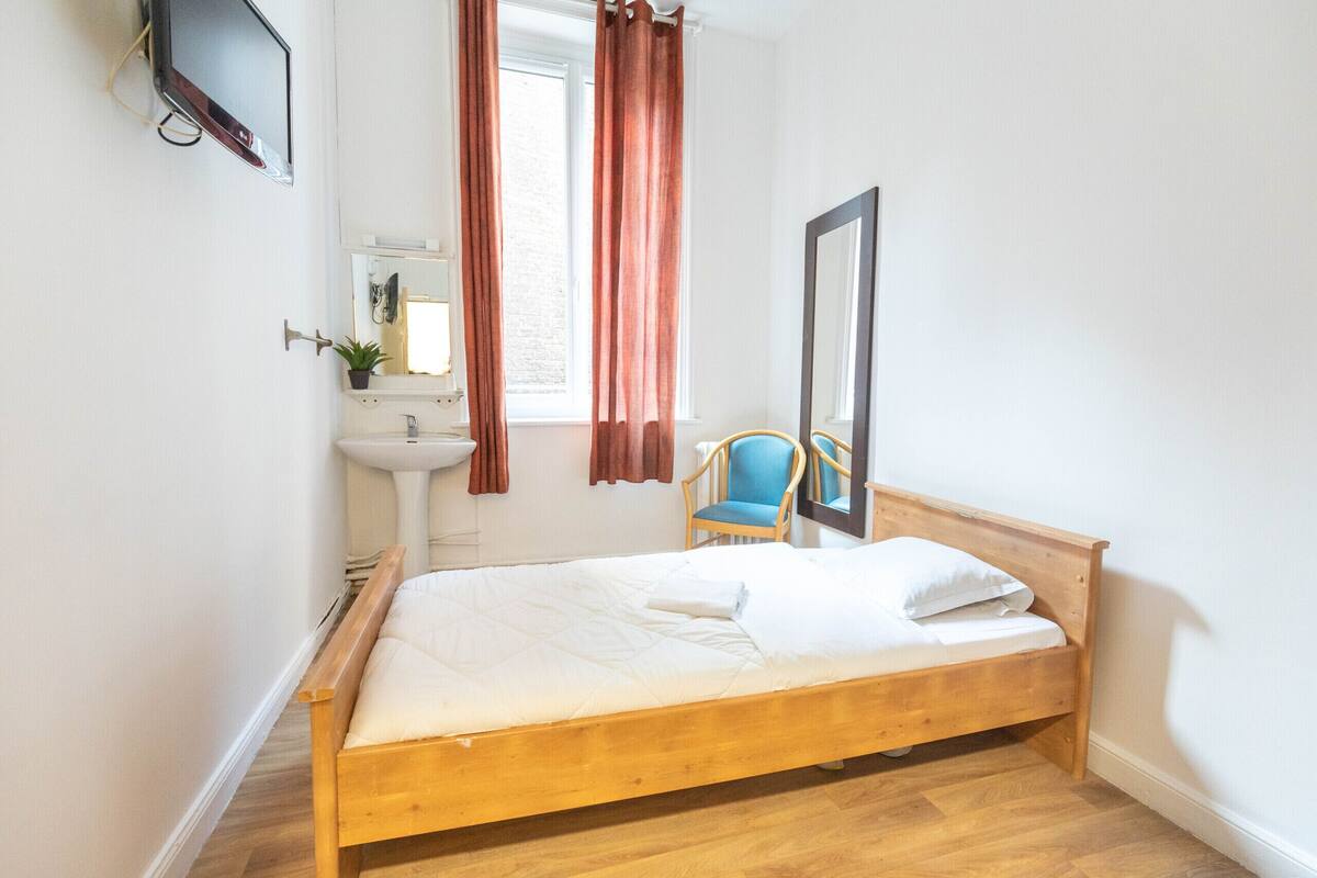Une chambre à coucher avec un lit en bois, une petite table de chevet, une chaise, un miroir et une fenêtre avec des rideaux.