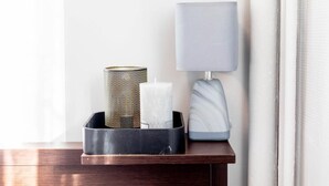 Ein Holznachttisch mit einer schwarzen Ablage, auf der sich eine dekorative Metallvase, eine weiße Kerze und eine Marmorlampe befinden.