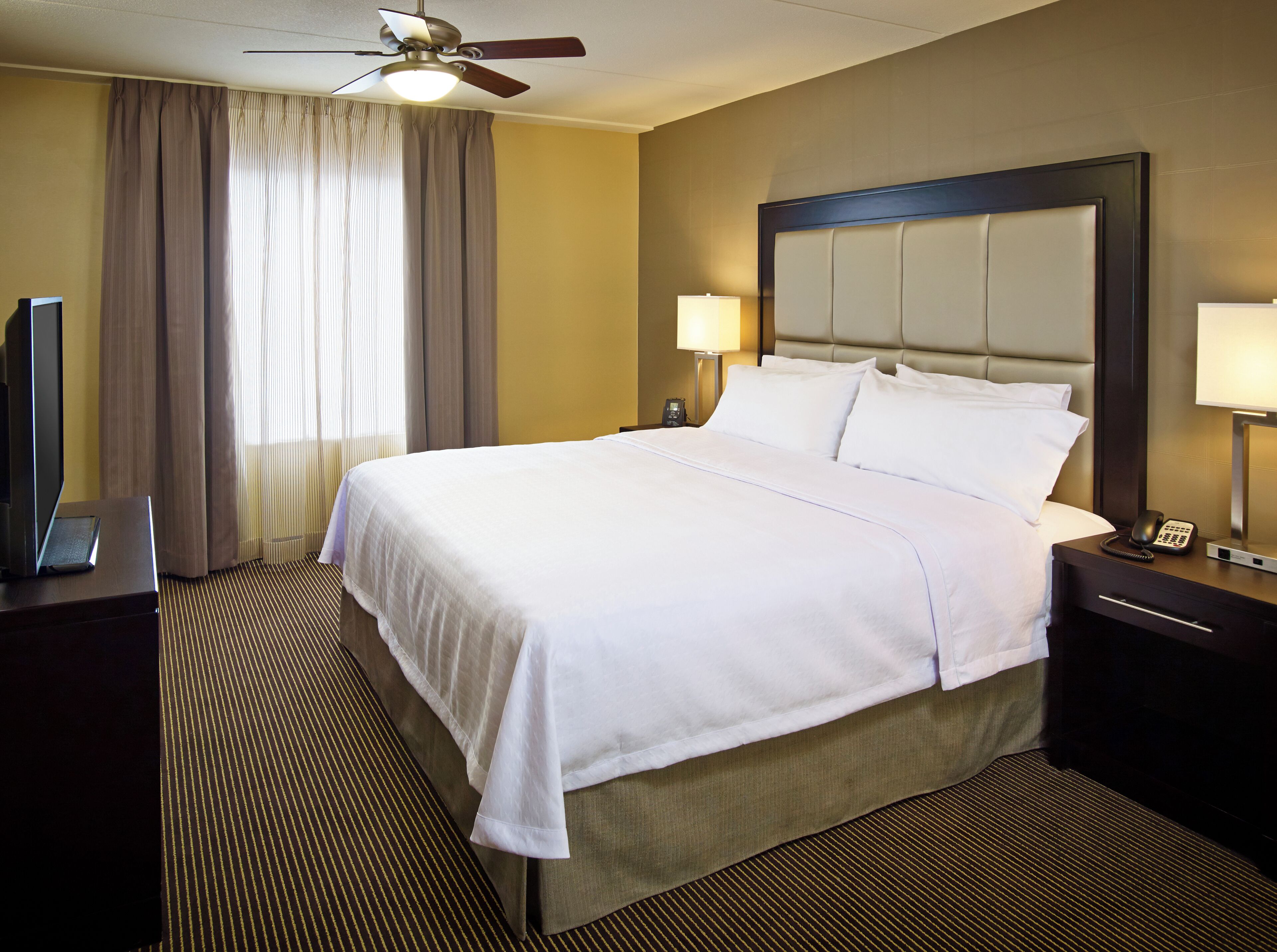 A hotel room with a large bed, a ceiling fan, a desk with a computer, a lamp, and a telephone.