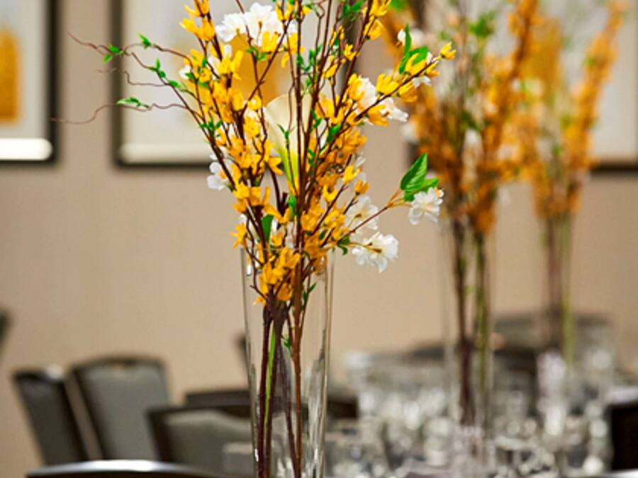 Una mesa de comedor con cubiertos de vidrio transparente y un centro de mesa con flores amarillas y blancas en un florero alto.