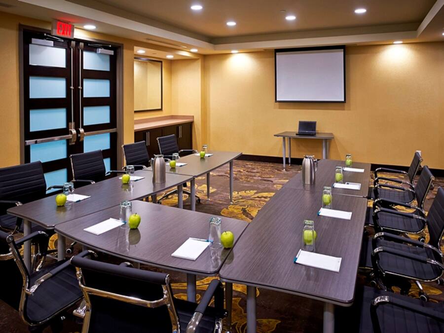 A conference room with long tables, chairs, a projector screen, and a small table with a laptop.