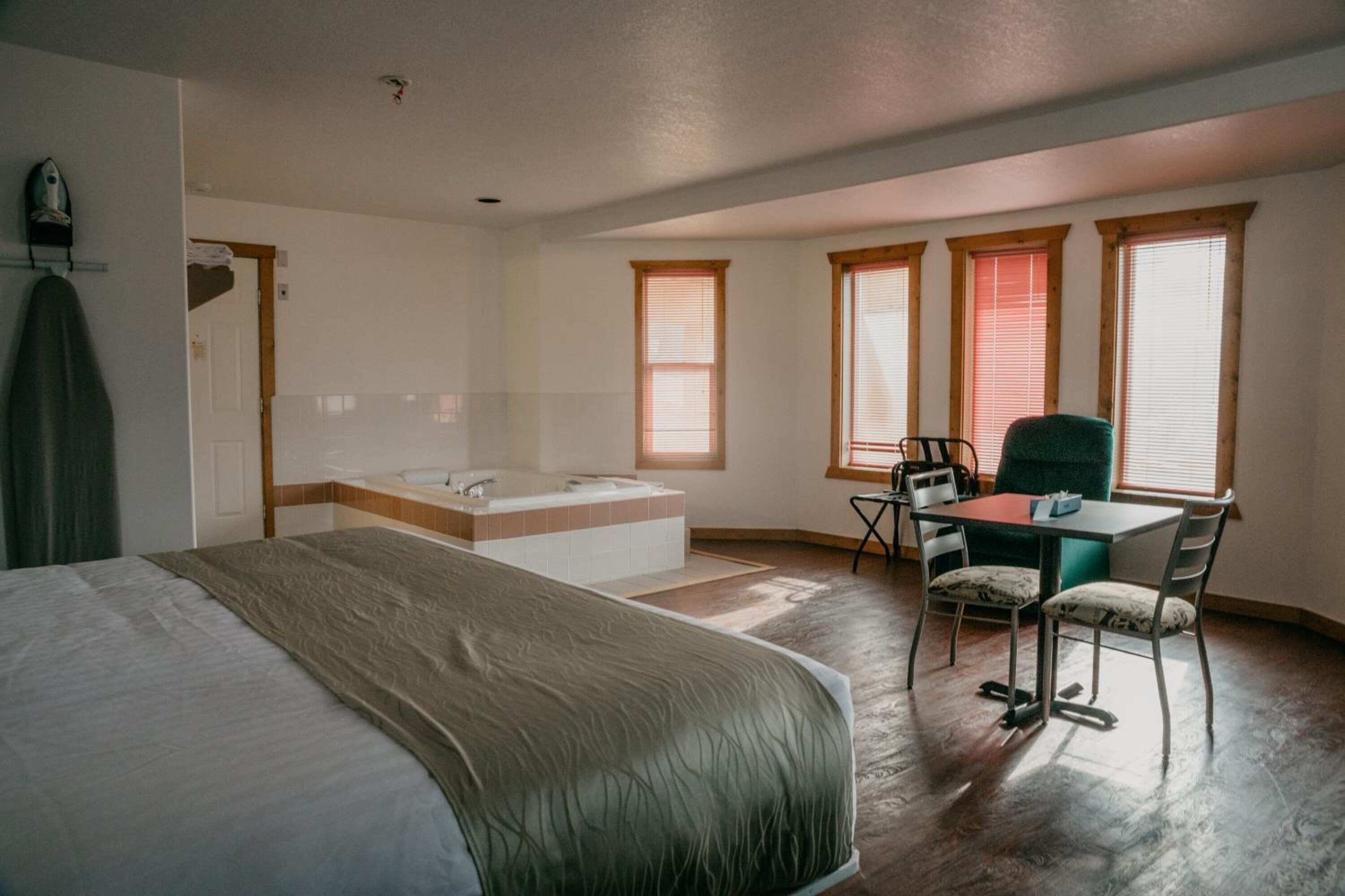 Habitación de hotel con una cama grande, un rincón de descanso con una mesa y sillas, una bañera y ventanas con persianas.