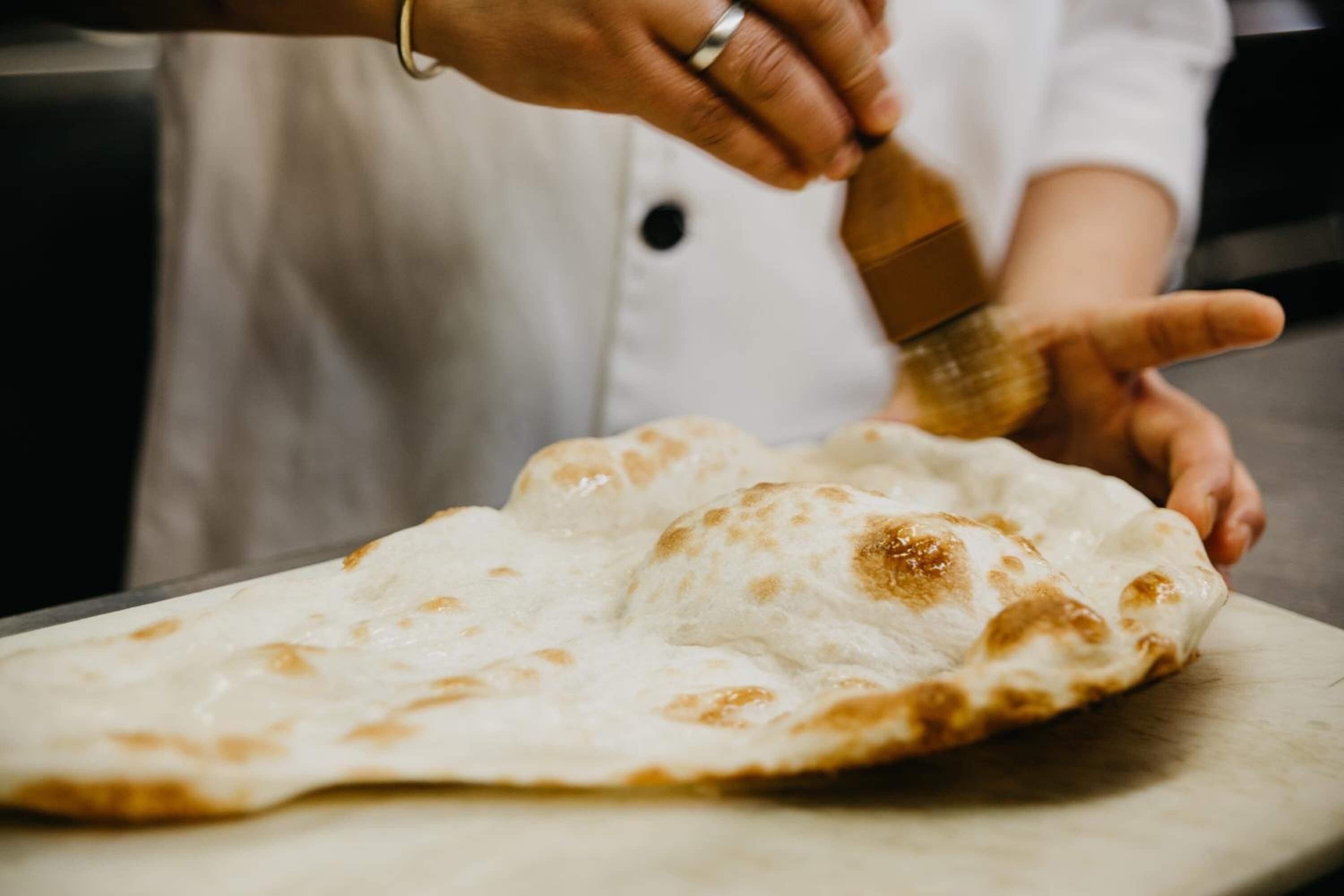 Une personne vêtue d’une blouse de chef blanche, tenant un rouleau à pâtisserie en bois au-dessus d’une grande pâte ronde et plate, posée sur une surface en bois.