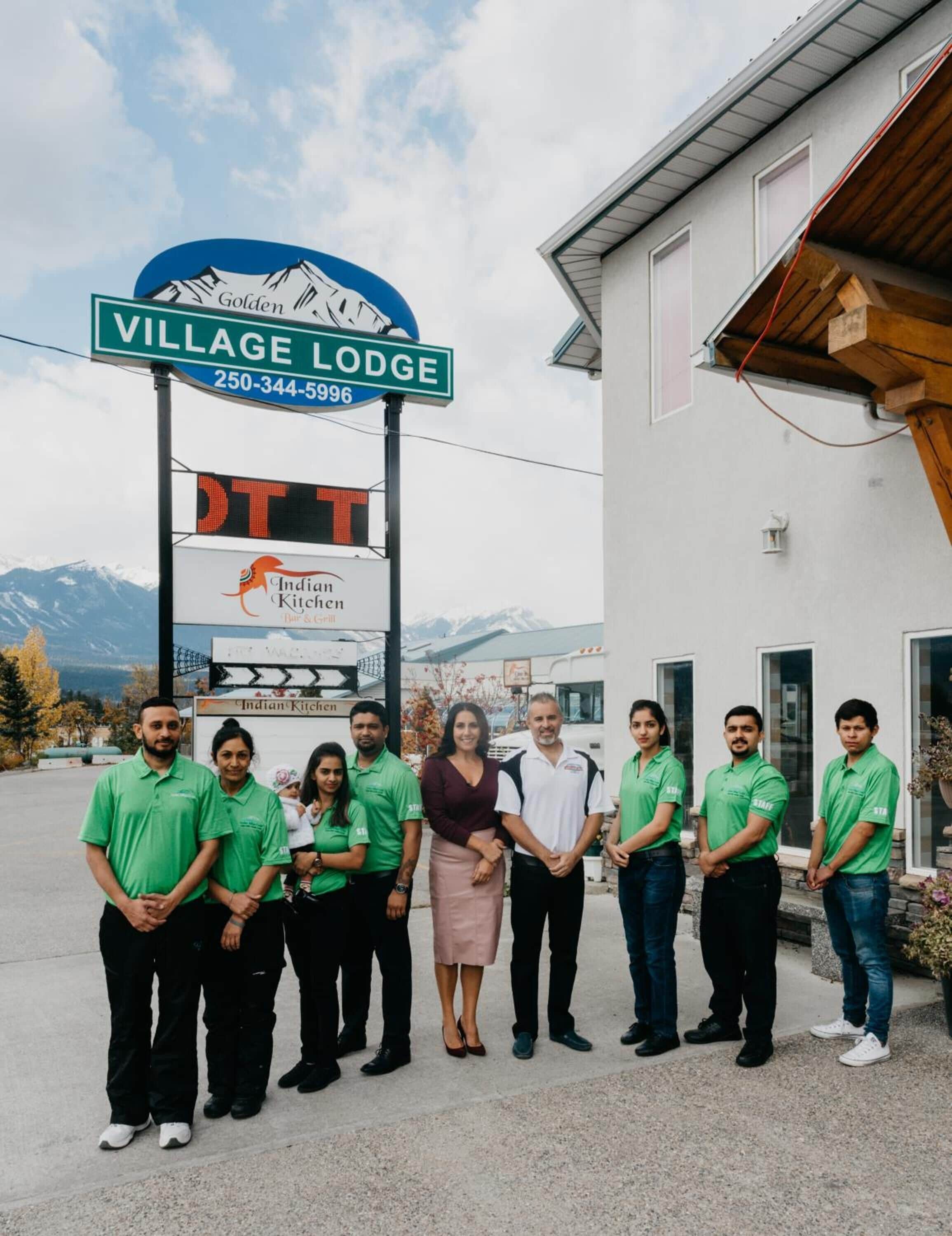 A group of people standing in front of a building with a sign that reads "Village Lodge" and a digital sign for "Indian Kitchen."