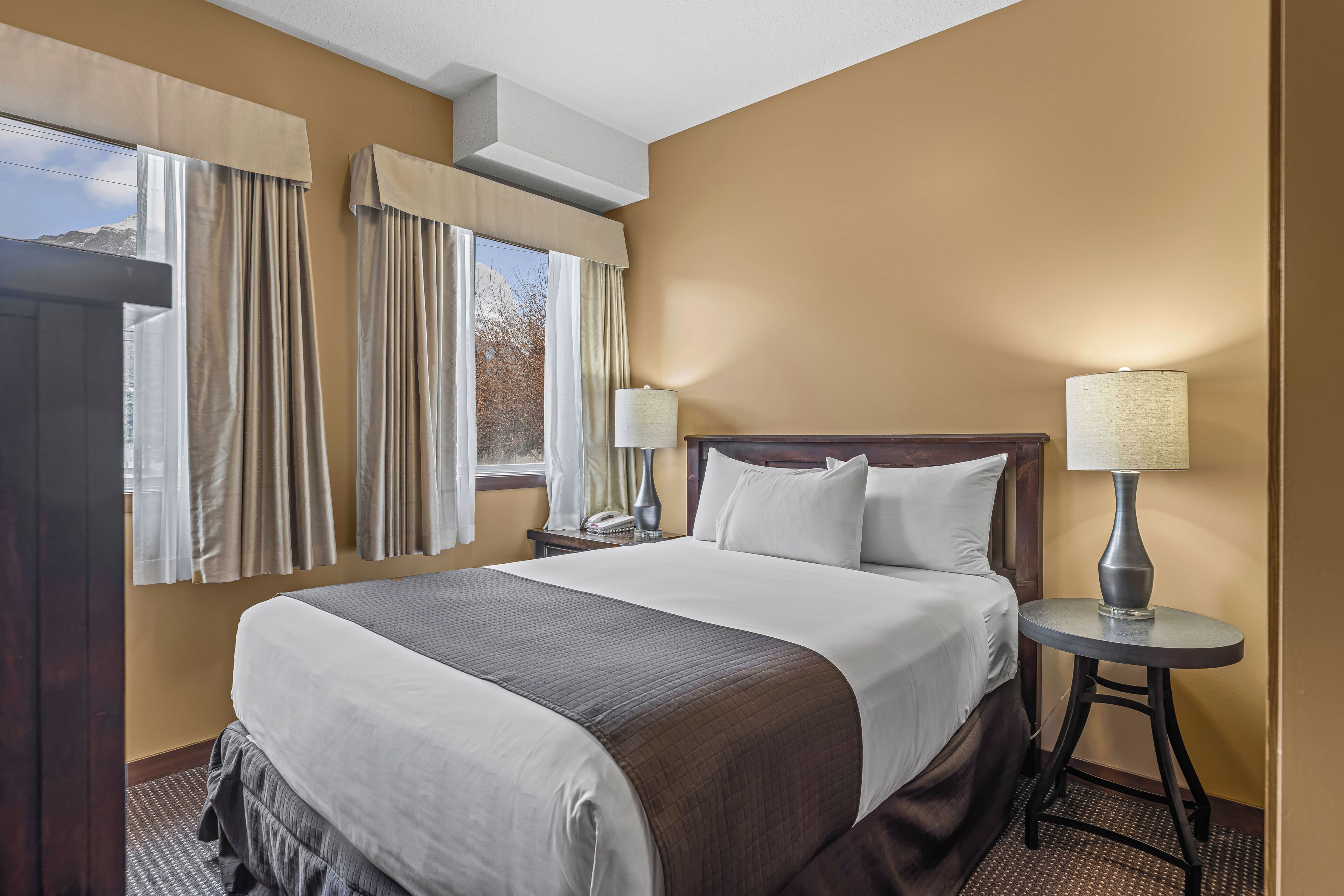 A hotel room with a large bed, two bedside tables with lamps, a window with curtains, and a wall-mounted air conditioner.