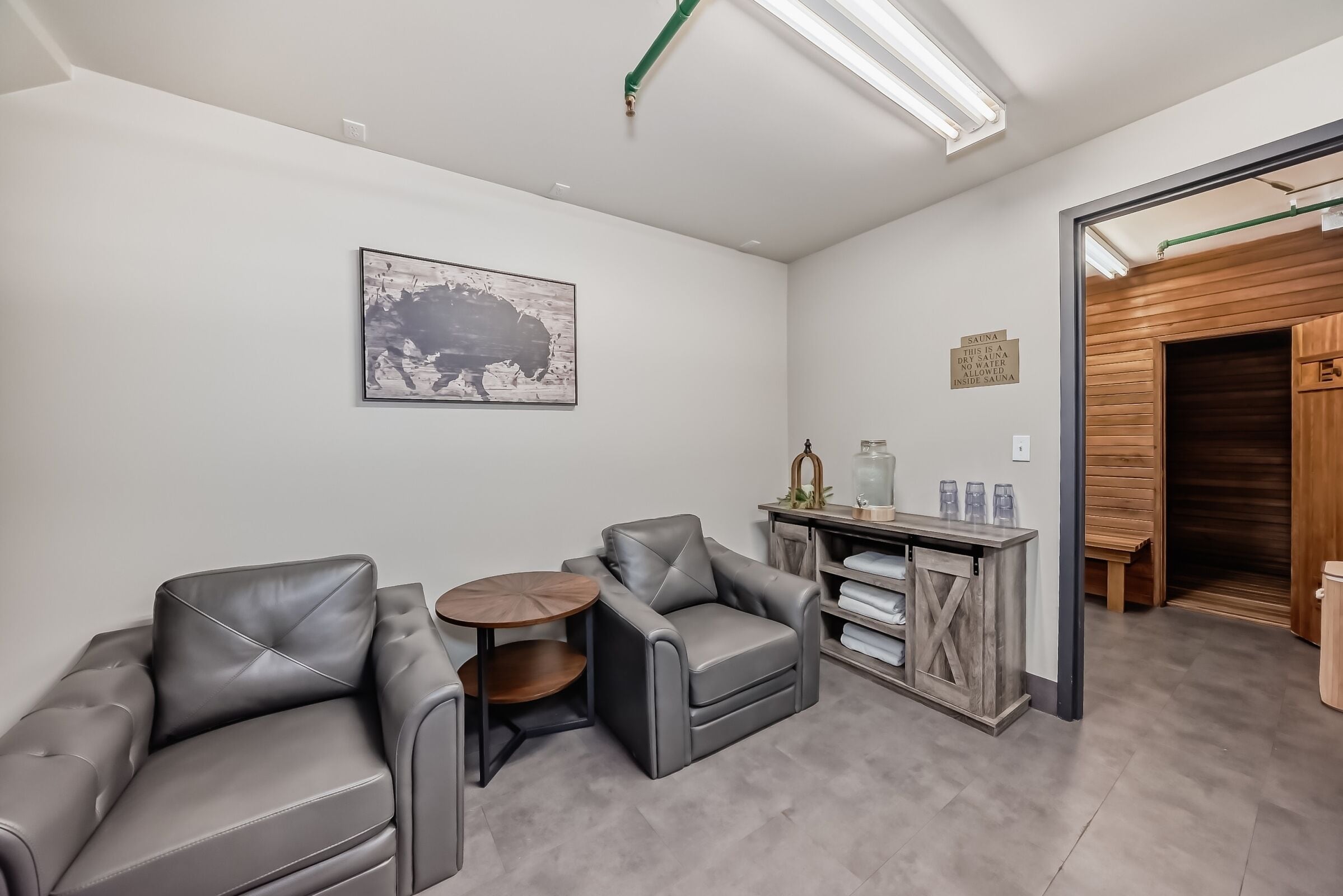 Une pièce moderne avec deux fauteuils en cuir, une petite table ronde, un meuble en bois et un panneau indiquant la présence d’un sauna.