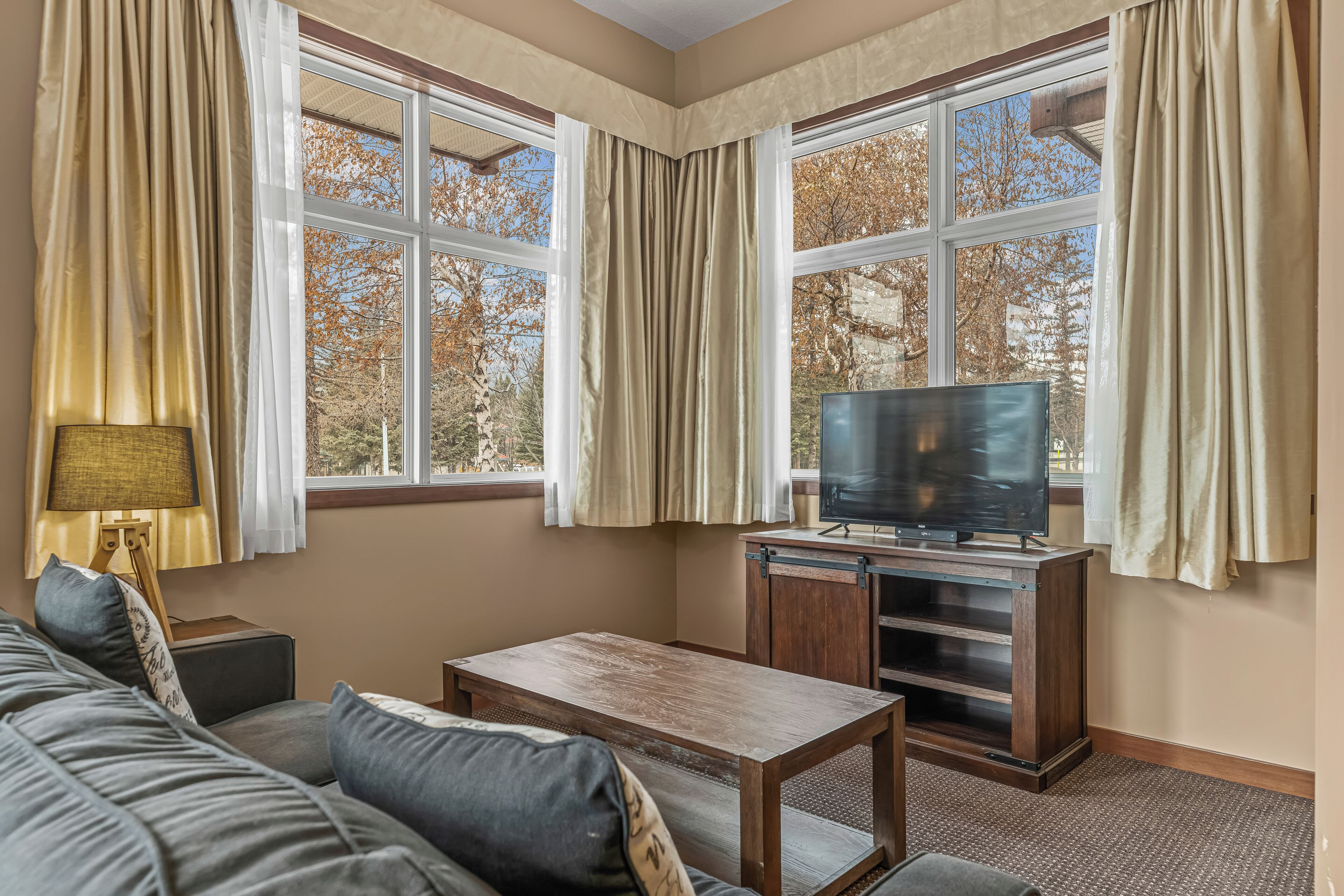 Ein Zimmer mit einer Couch, einem Holztisch, einem Fernseher auf einem Schrank und großen Fenstern mit Vorhängen.