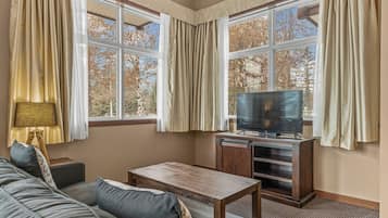A room with a sofa, a wooden table, a television on a stand, and large windows with curtains.