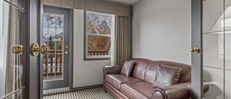 A room with a brown leather sofa, a patterned carpet, a large window with a view of trees and mountains, and a door with a mirror and curtains.