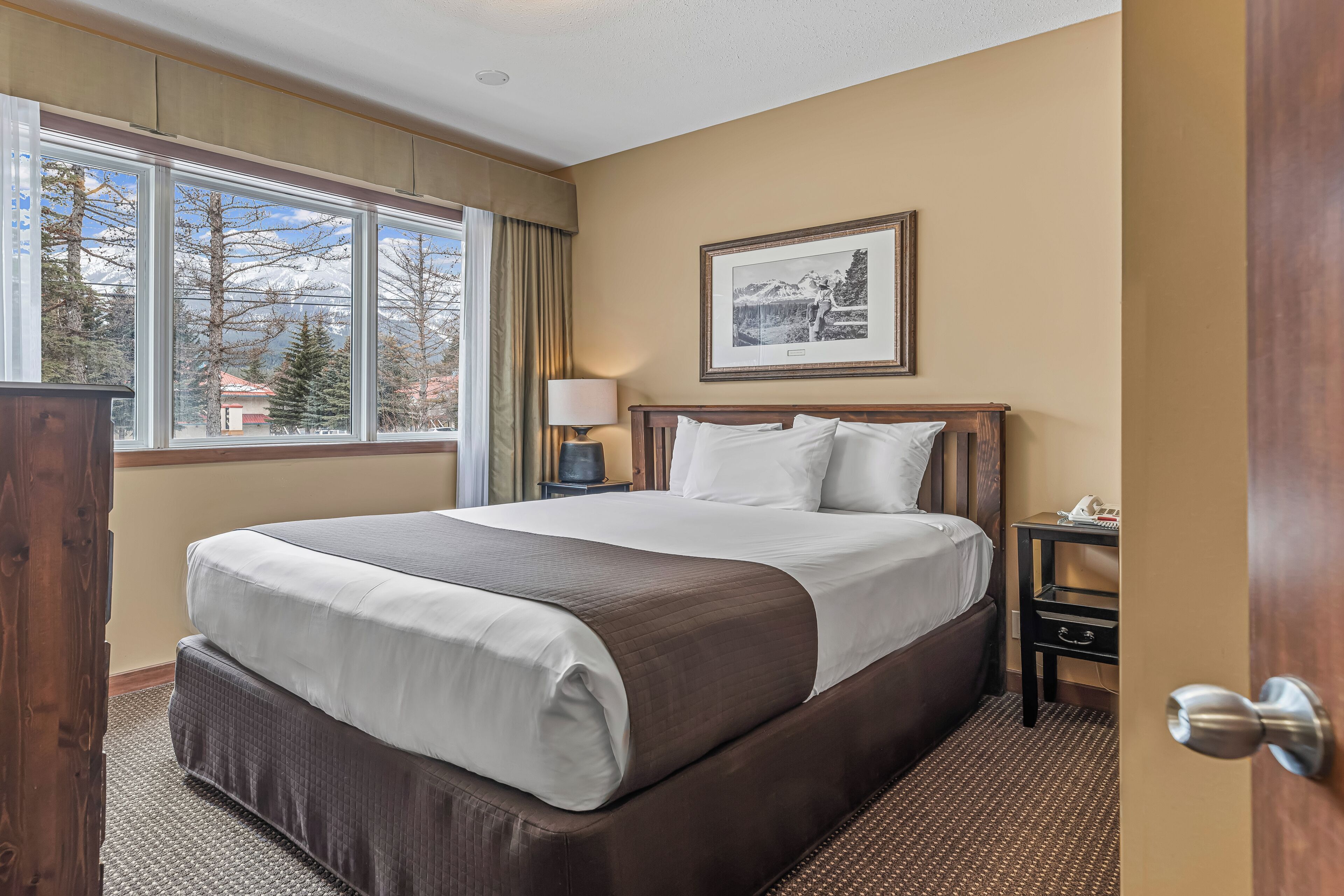 Quarto de hotel com uma cama grande, uma mesa de cabeceira, uma luminária, um quadro na parede e uma janela com vista para árvores e um edifício.