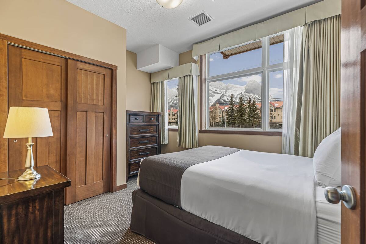 A hotel room with a bed, a dresser, a lamp, a window with curtains, and a view of mountains.