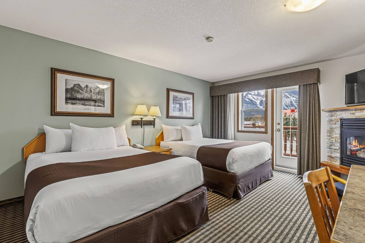A hotel room with two beds, a fireplace, a dining area, and a view of mountains.