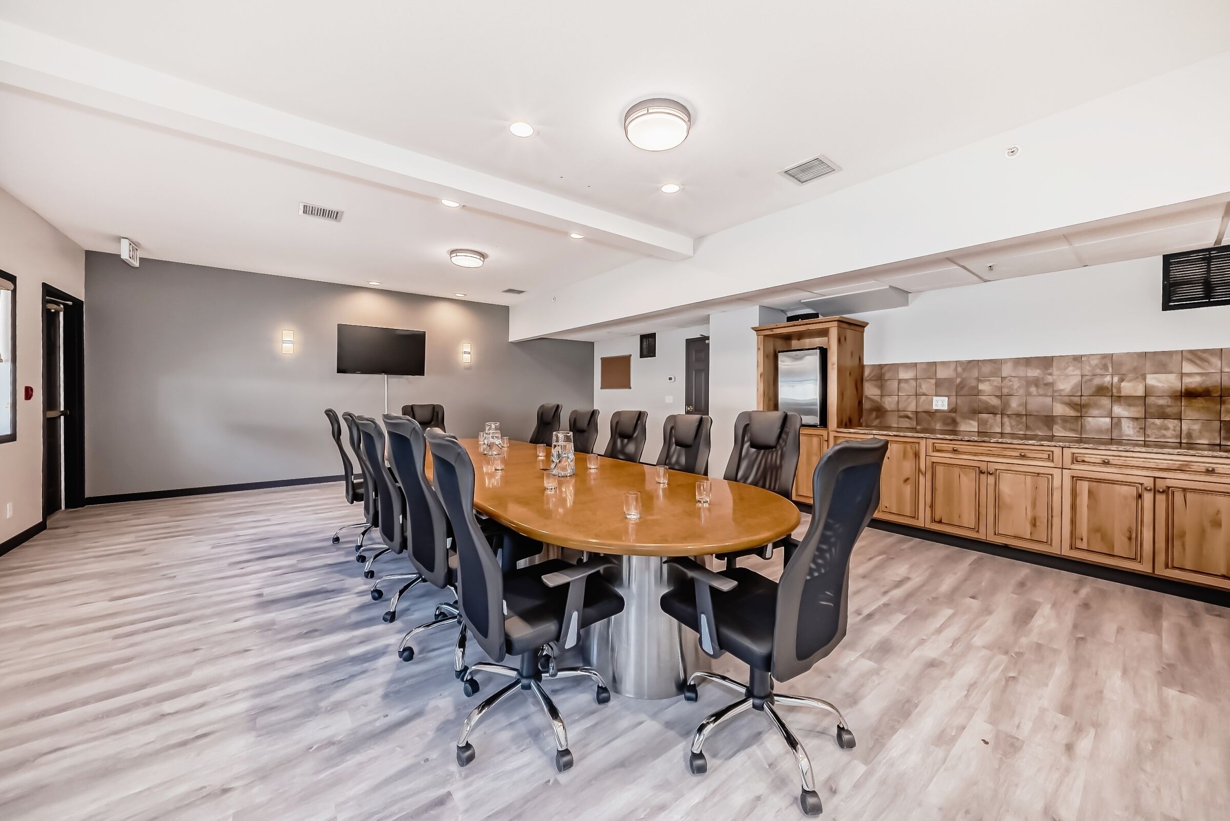 A conference room with a large oval table, black chairs, a flat-screen TV, and a wooden cabinet.