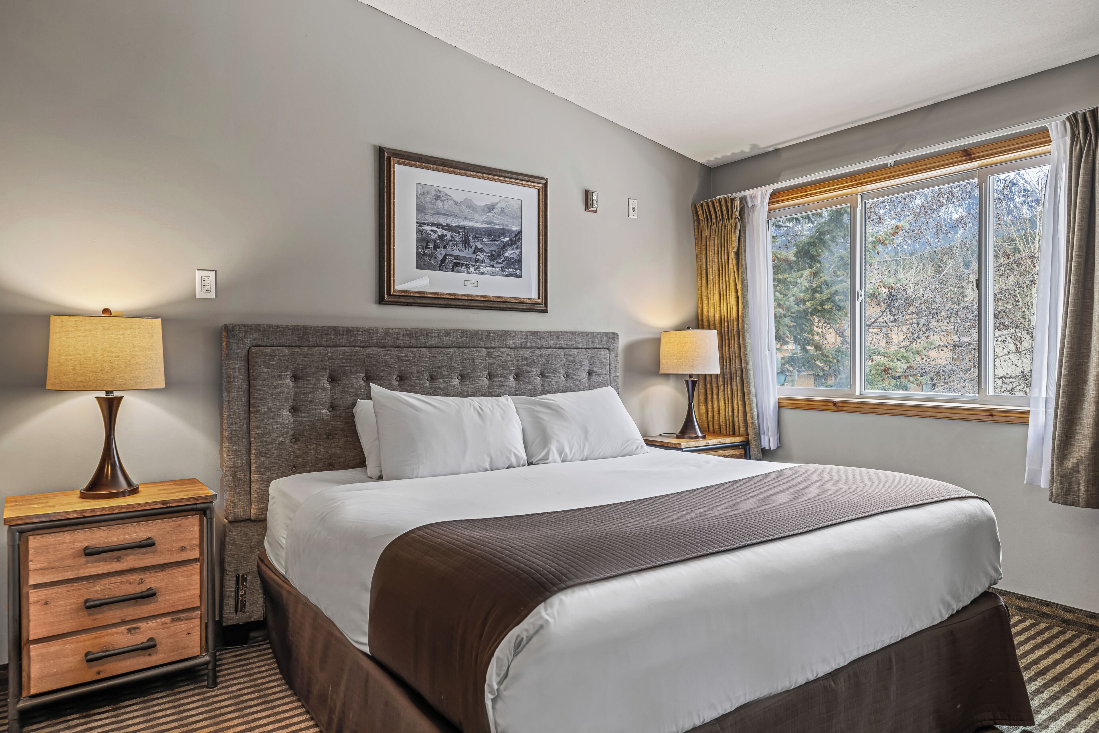 Habitación de hotel ordenada con una cama grande, dos mesitas de noche, un cuadro en la pared y una ventana con cortinas.