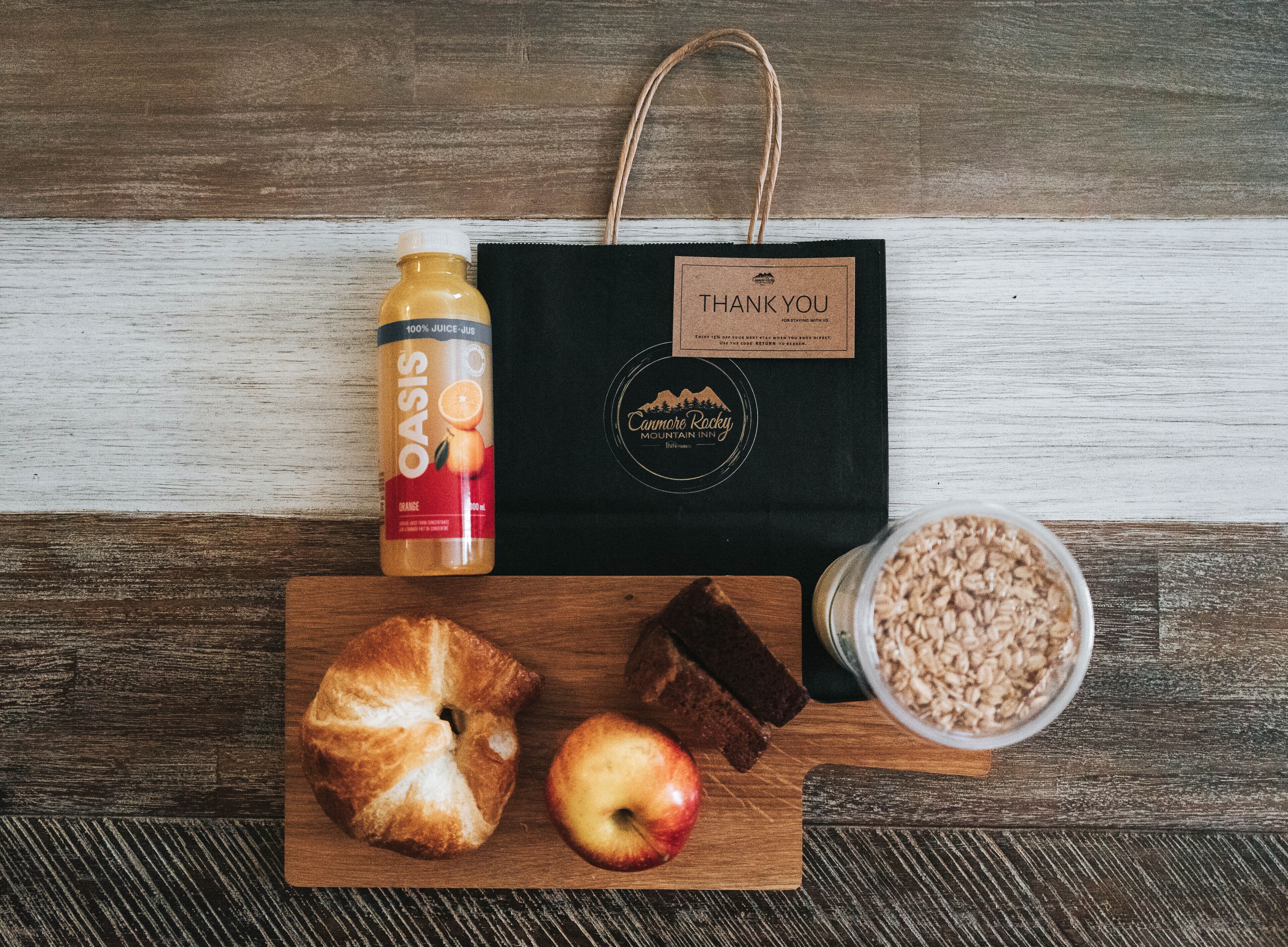 A wooden table with a bottle of orange juice, a bag with a "THANK YOU" label, a croissant, an apple, chocolate pieces, and a bowl of cereal.