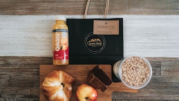 A wooden table with a bottle of orange juice, a bag with a "THANK YOU" label, a croissant, an apple, chocolate pieces, and a bowl of cereal.