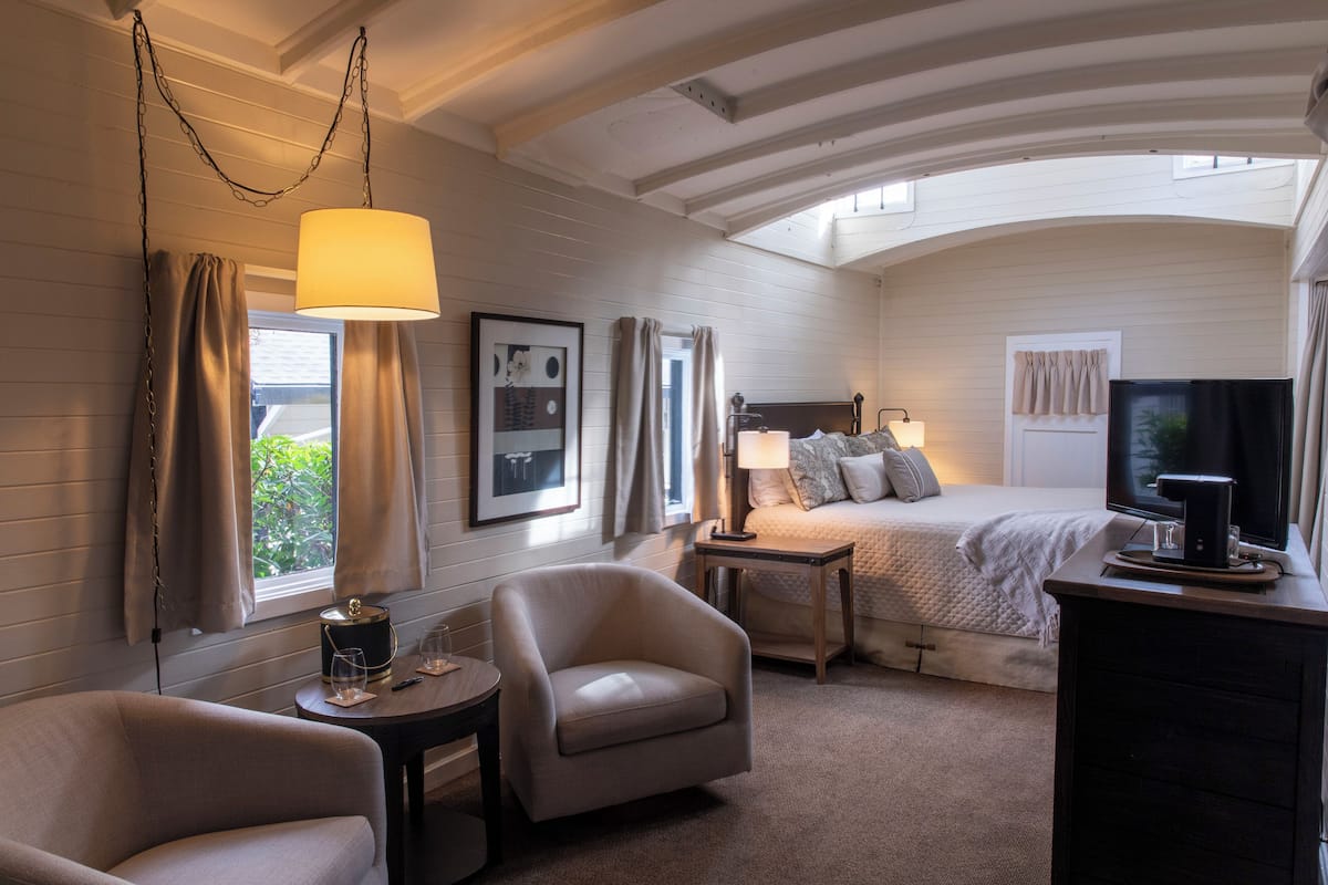 A hotel room with a bed, two armchairs, a bedside table with a lamp, a TV, and a window with curtains.